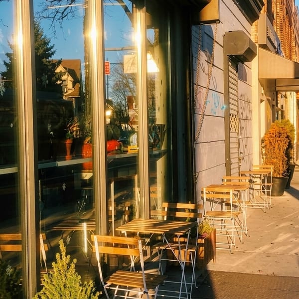 Sunny street view of a cafe with outdoor seating, reflecting the street and trees.