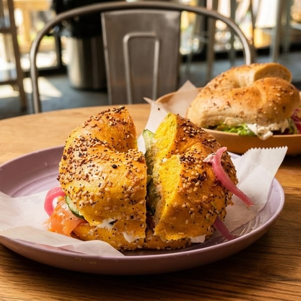 Two bagels on a wooden table, one with salmon and cream cheese, the other with greens.
