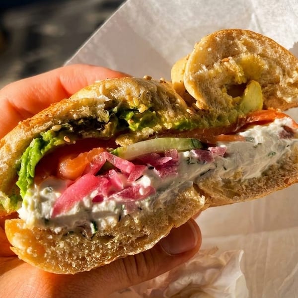 A hand holds a sliced bagel sandwich filled with cream cheese, salmon, avocado, and pickled onions.