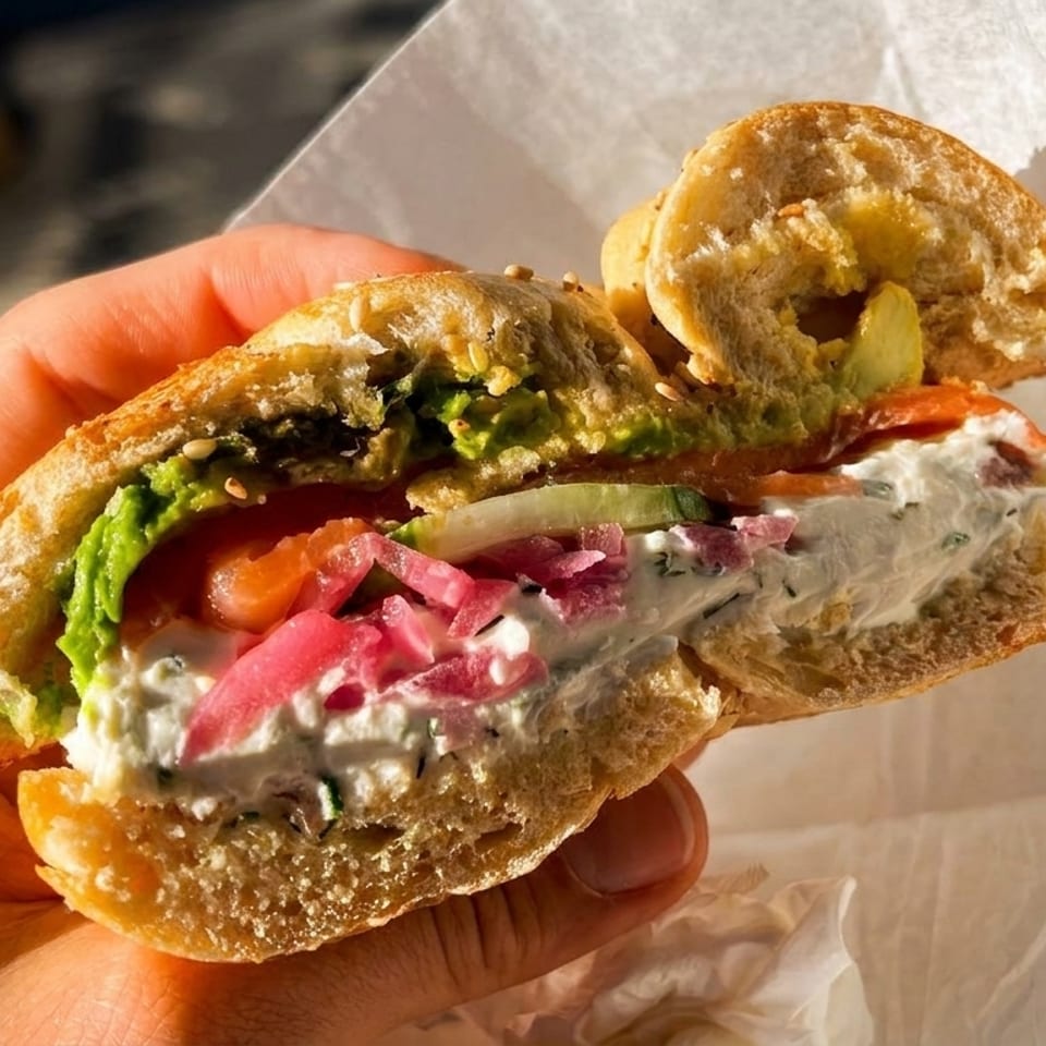 A hand holds a sliced bagel sandwich filled with cream cheese, salmon, avocado, and pickled onions.