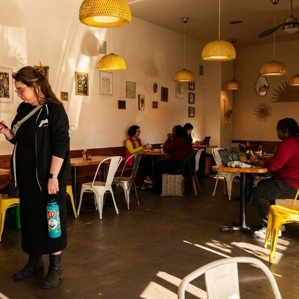 A woman stands in a sunlit cafe, checking her phone, while other patrons sit at tables.