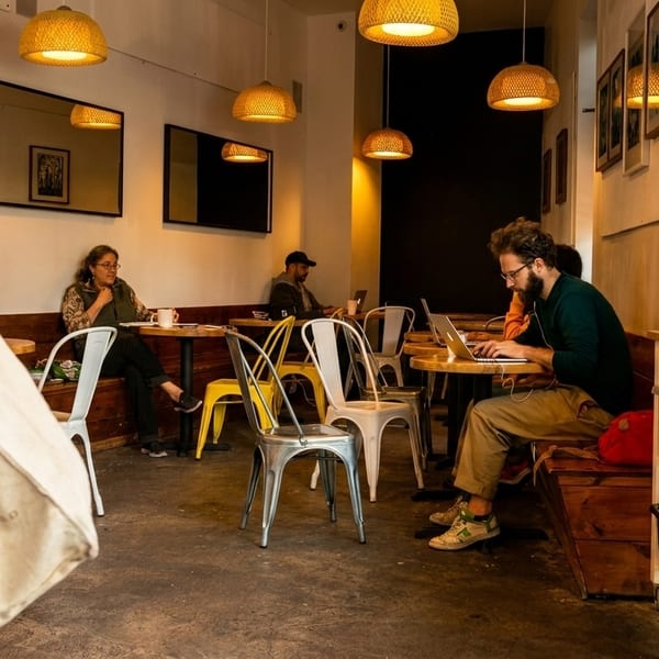 Customers work on laptops and relax at tables in a cozy, brightly lit cafe with woven pendant lights.