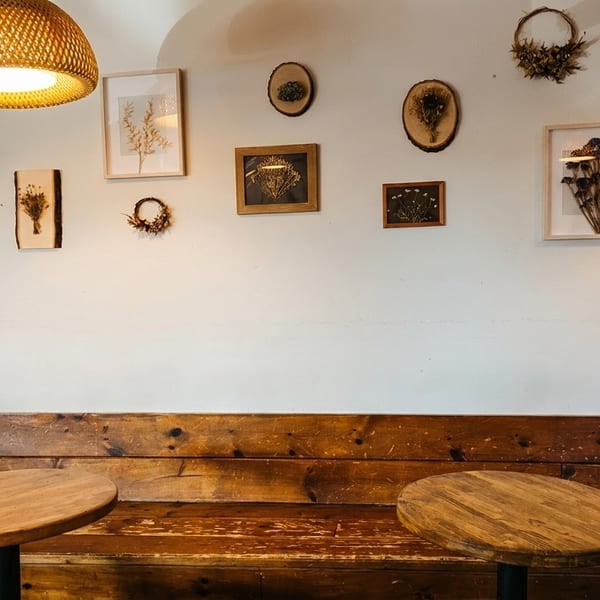 A cozy cafe interior with wooden tables, a bench, and framed dried floral art on a white wall.