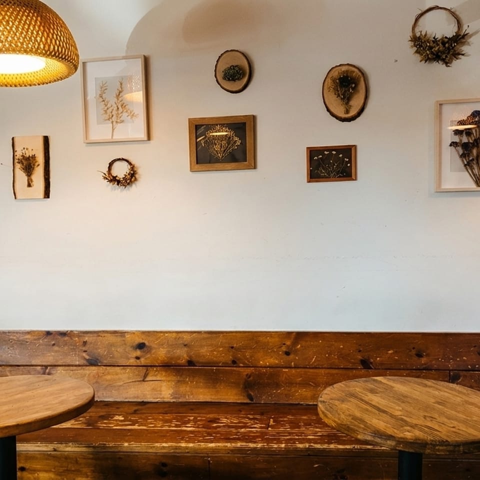 A cozy cafe interior with wooden tables, a bench, and framed dried floral art on a white wall.