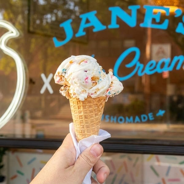 A hand holds a waffle cone with scoops of colorful ice cream in front of Jane's Cream shop.