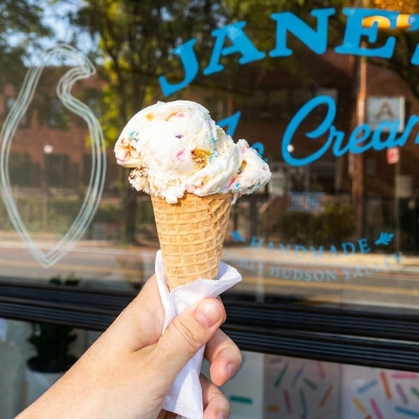 A hand holds a waffle cone with scoops of colorful ice cream in front of Jane's Ice Cream.